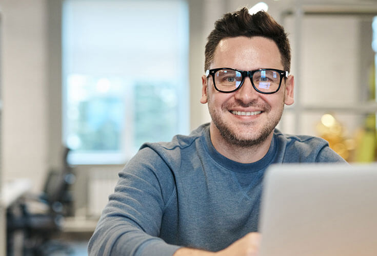 A male student with glasses working on his laptop while looking at the camera